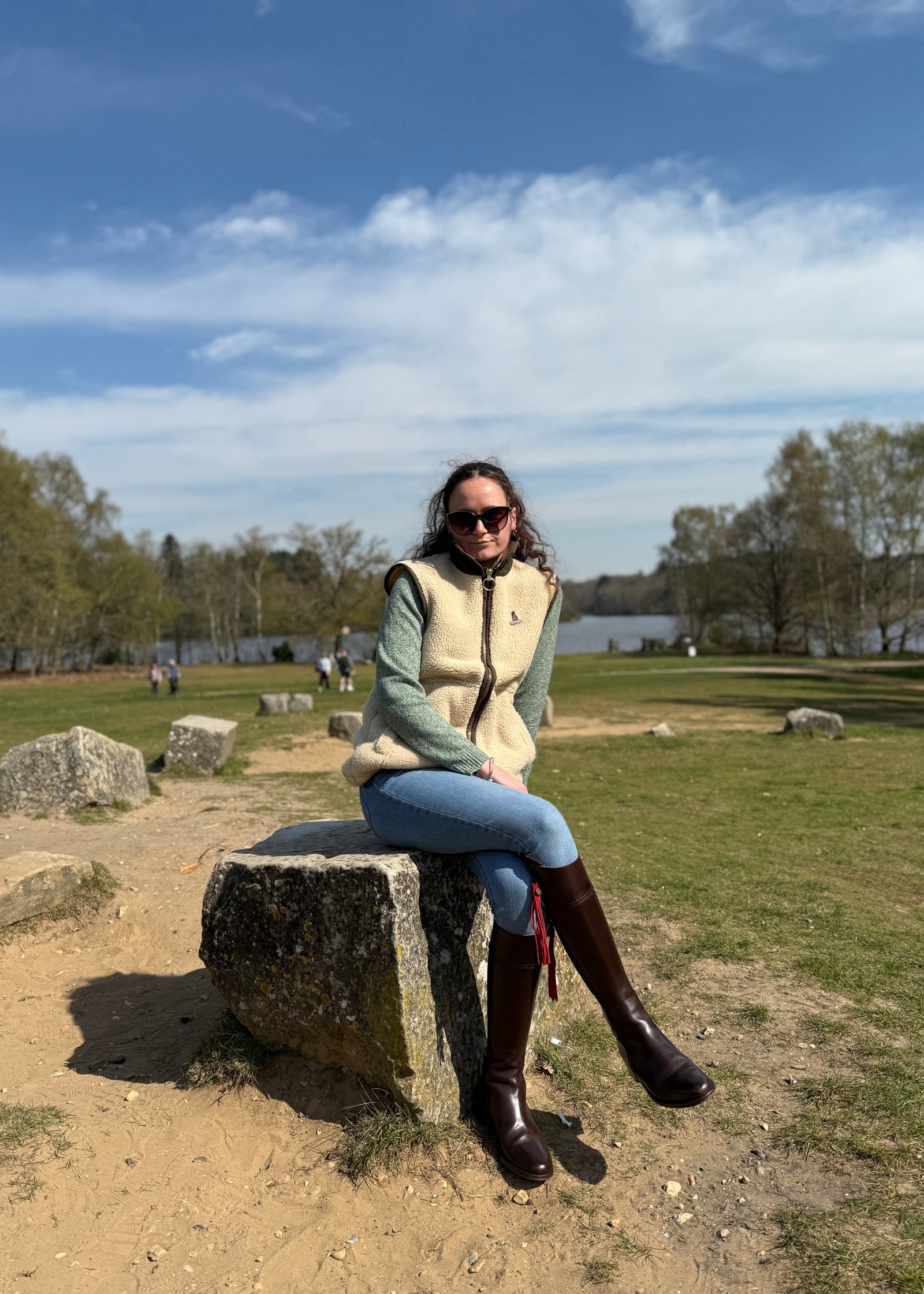 woman sitting in a park with a lake in the background whilst wearing boots, jeans and a beige gilet
