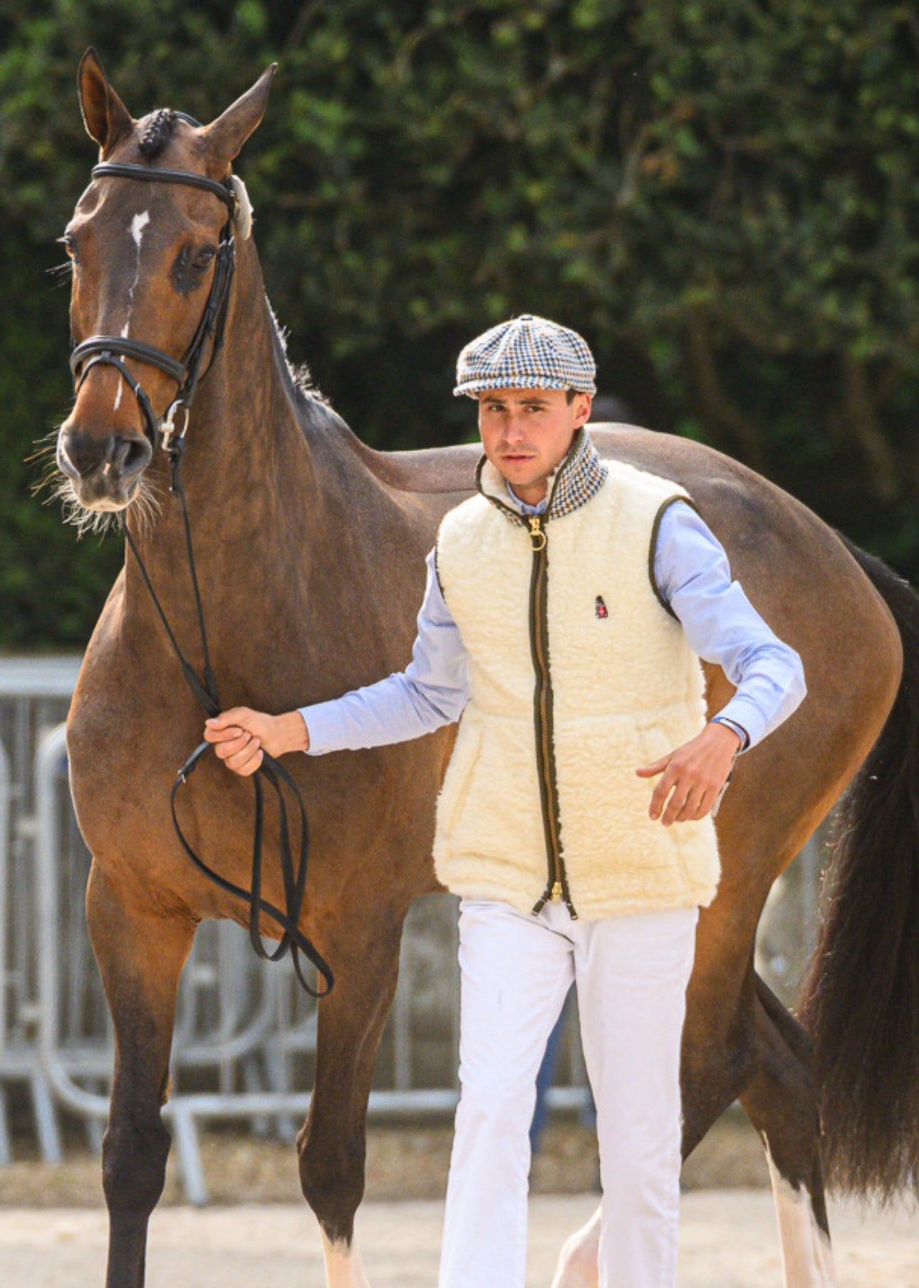 Person in equestrian attire standing next to a horse on a gravel path with greenery in the background