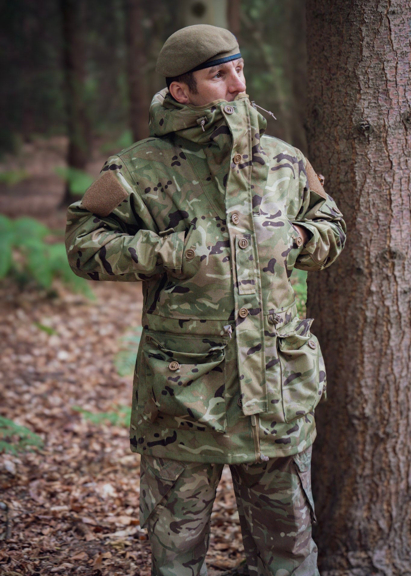 Person in camouflage clothing standing next to a tree in a forest setting