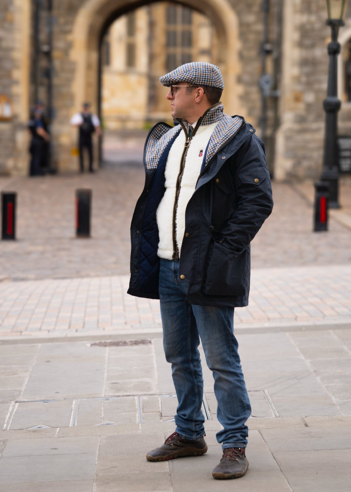 Man wearing a coat, scarf, and flat cap standing in front of a stone building.