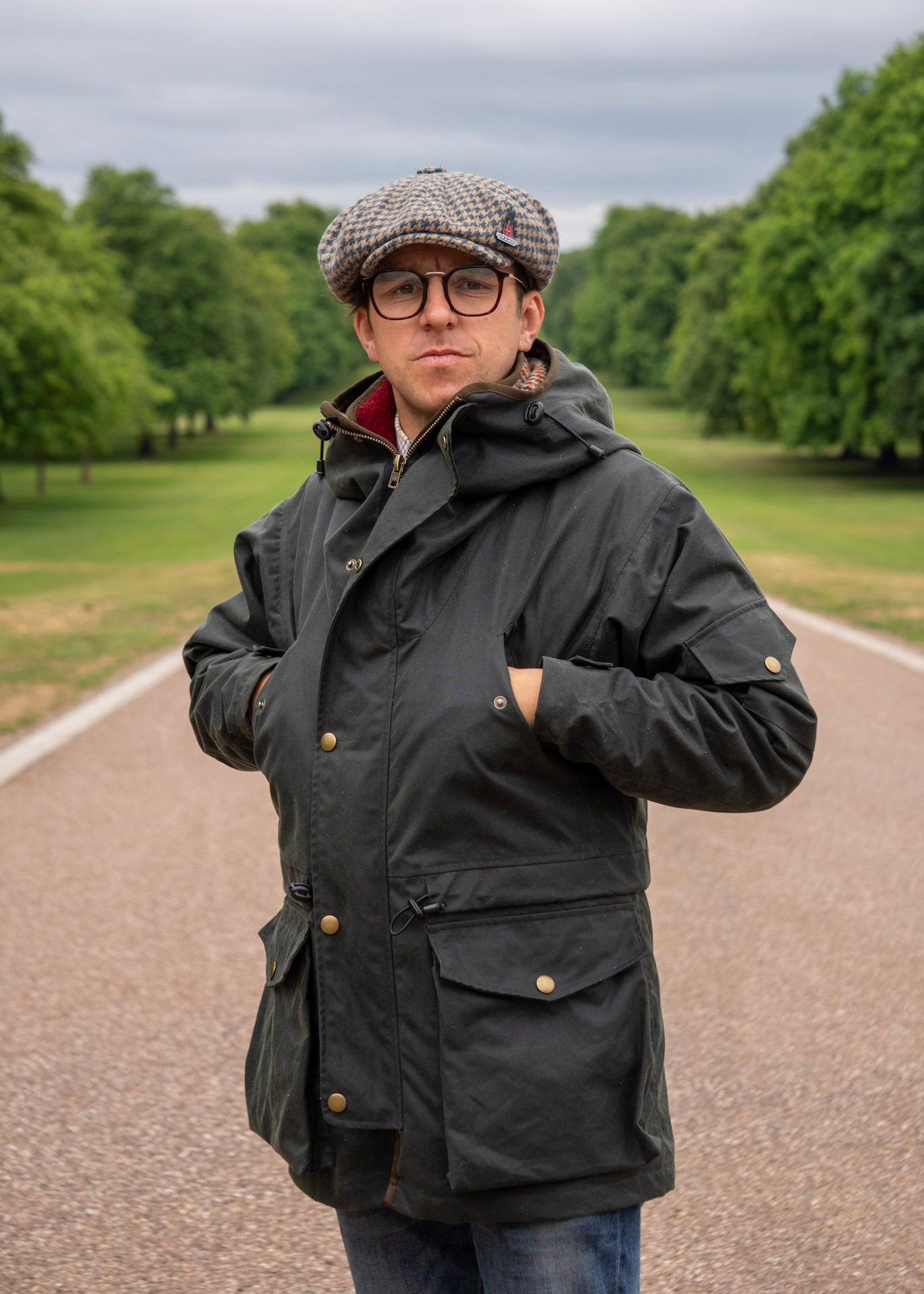 Man wearing a dark wax jacket and checkered cap standing on a path with trees in the background