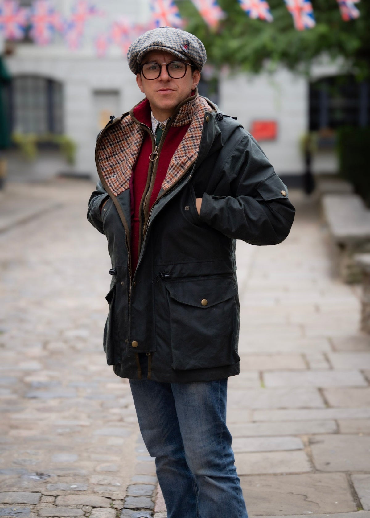 Man wearing a wax jacket, red gilet, and cap on a street with British flags in the background