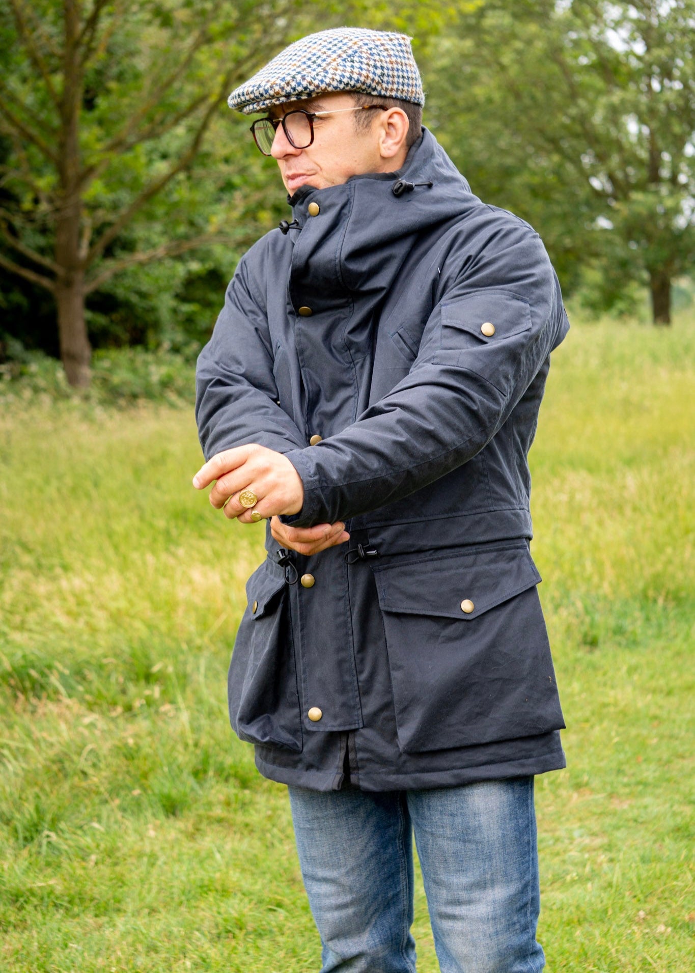 Man wearing a dark wax coat, jeans, and a cap standing in a grassy field.