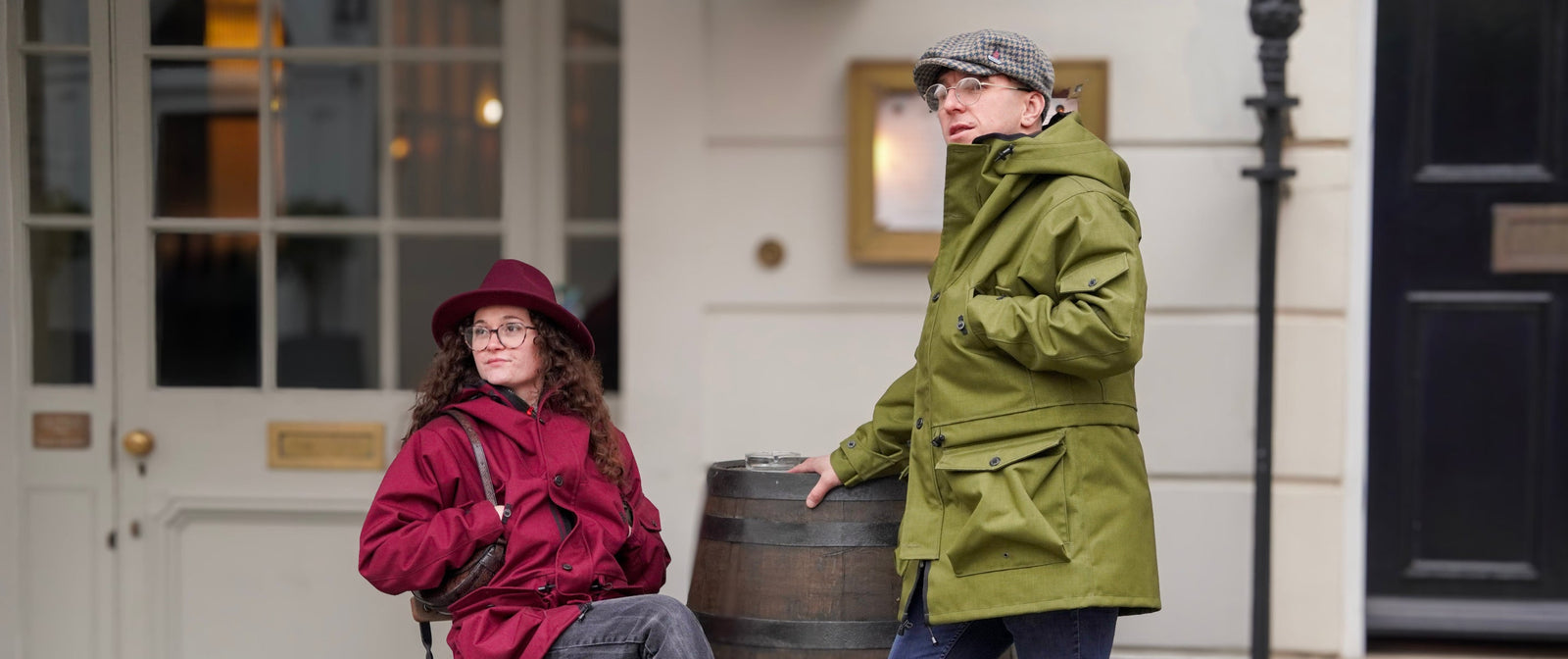 Two people in raincoats standing next to a barrel on a city street.