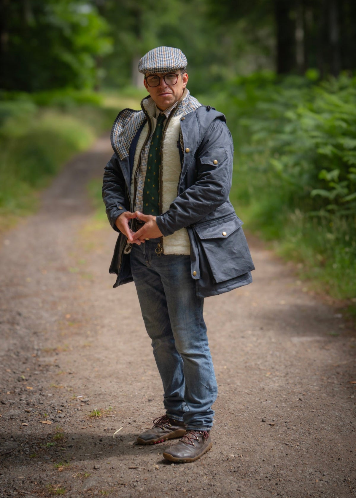 Man standing on a path in a forest wearing a way jacket, wool gilet, and hat.