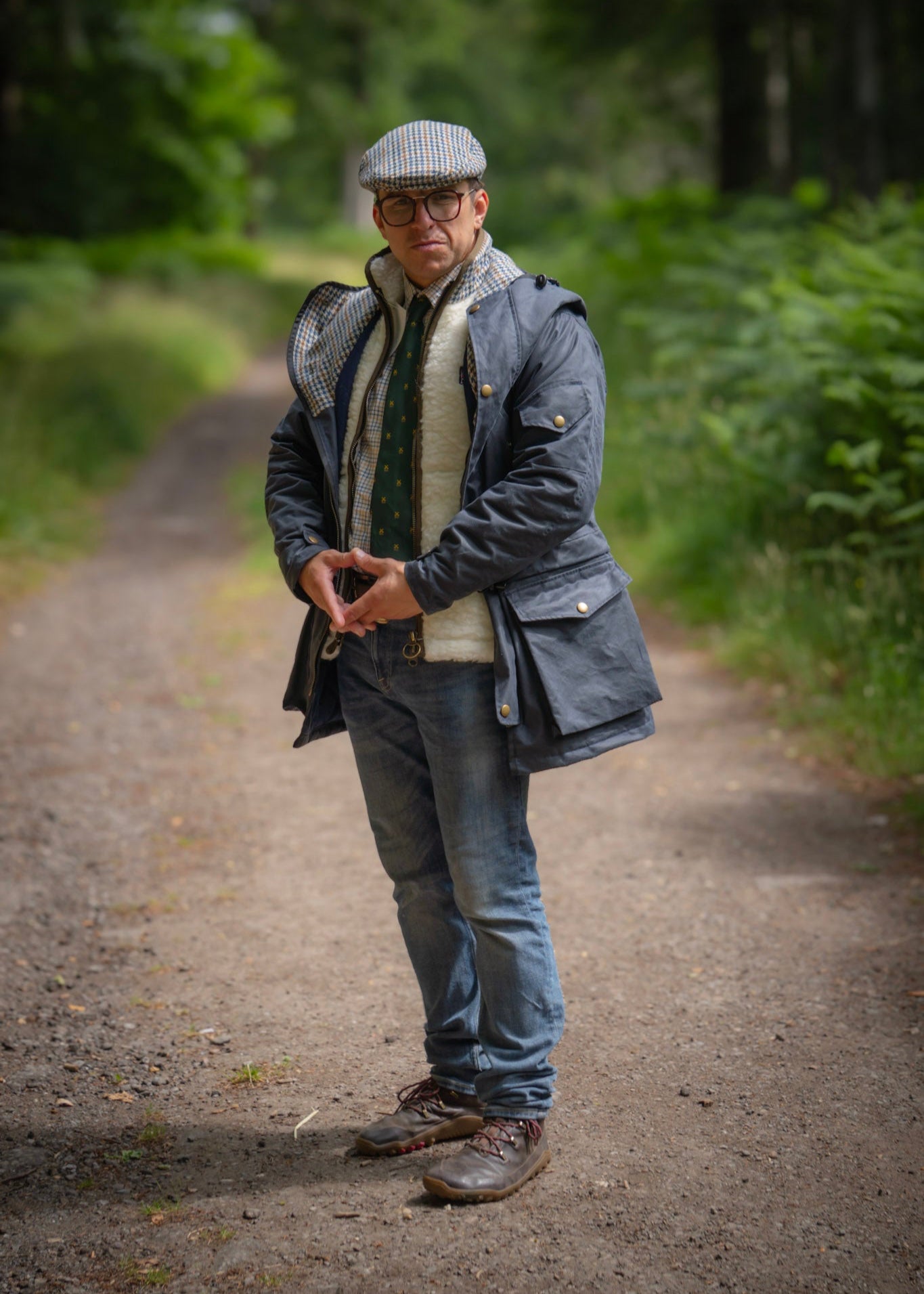 Man standing on a path in a forest wearing a way jacket, wool gilet, and hat.