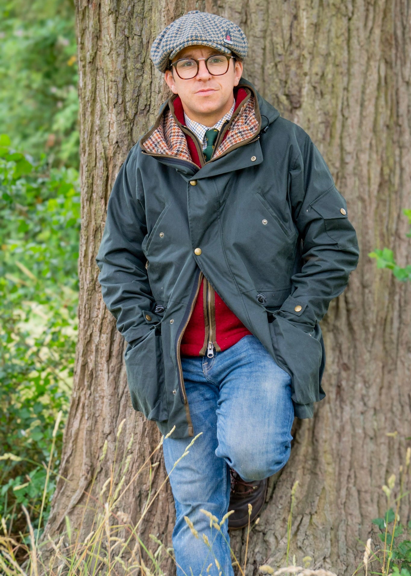 Man wearing a dark wax jacket, red gilet, and gray cap standing against a tree in a forest setting.