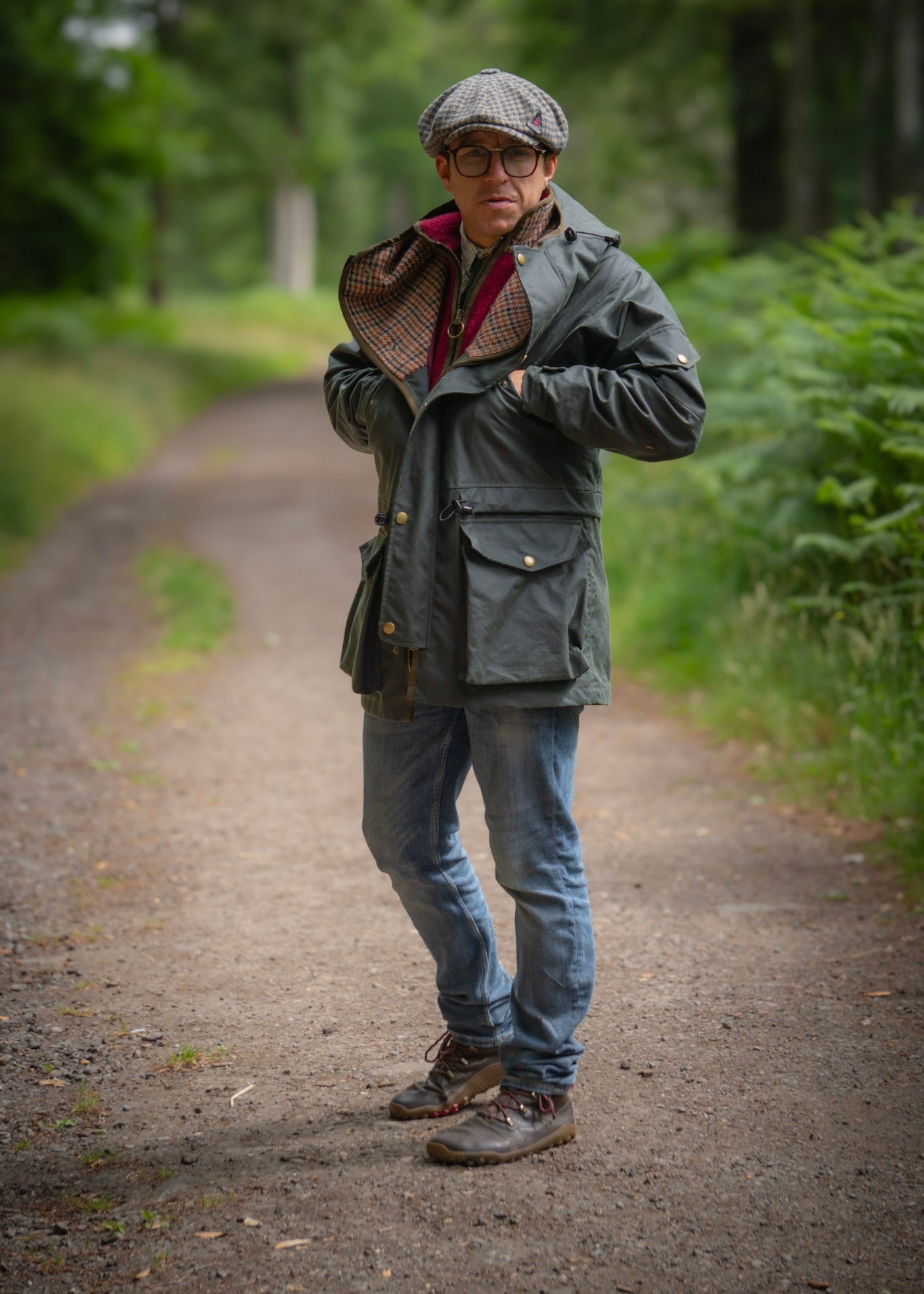 Person wearing a green wax jacket, cap, and glasses standing on a path in a forest.
