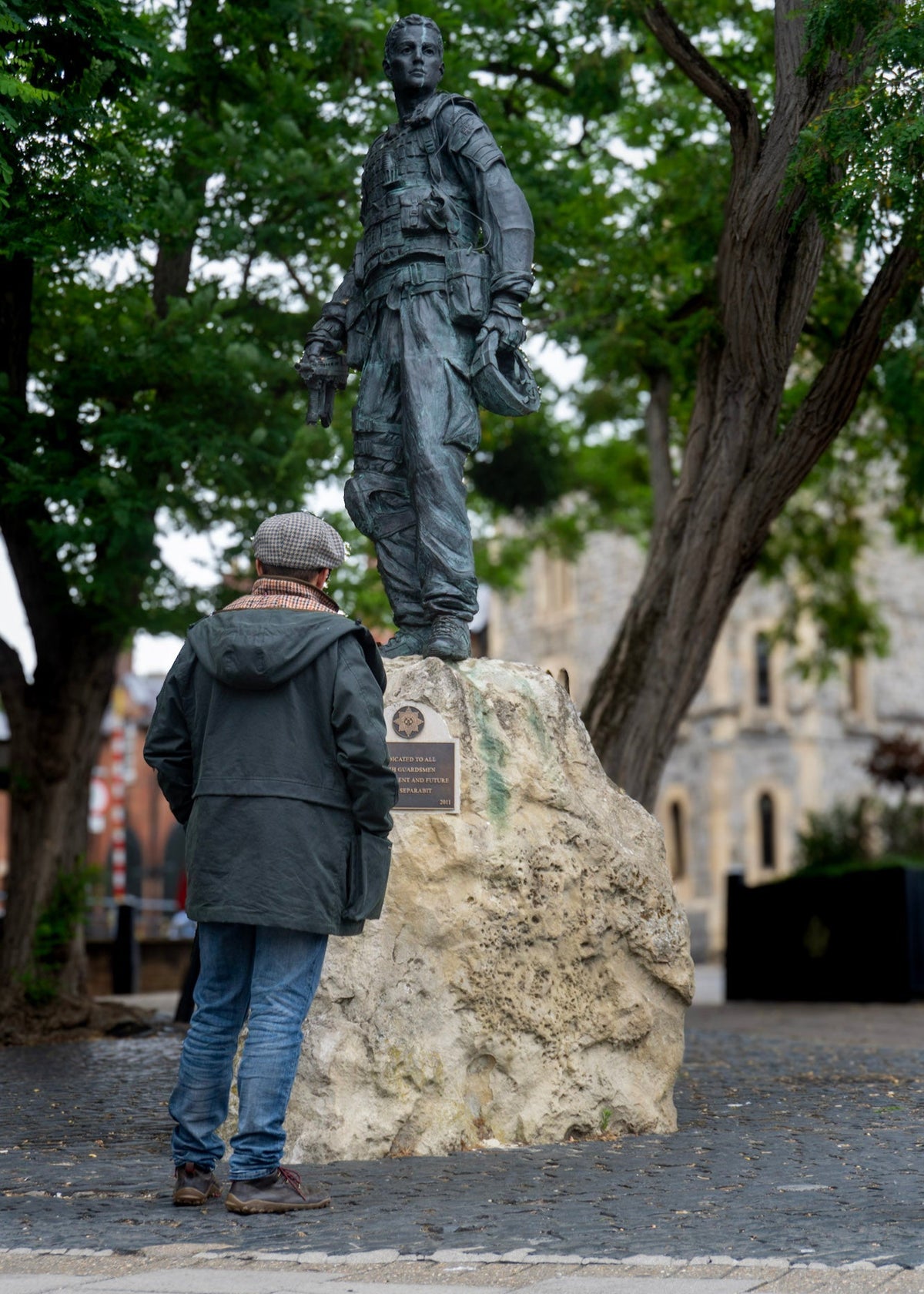 Person looking at a statue of a soldier on a rock with trees and building in the background, wearing a wax jacket and flat cap