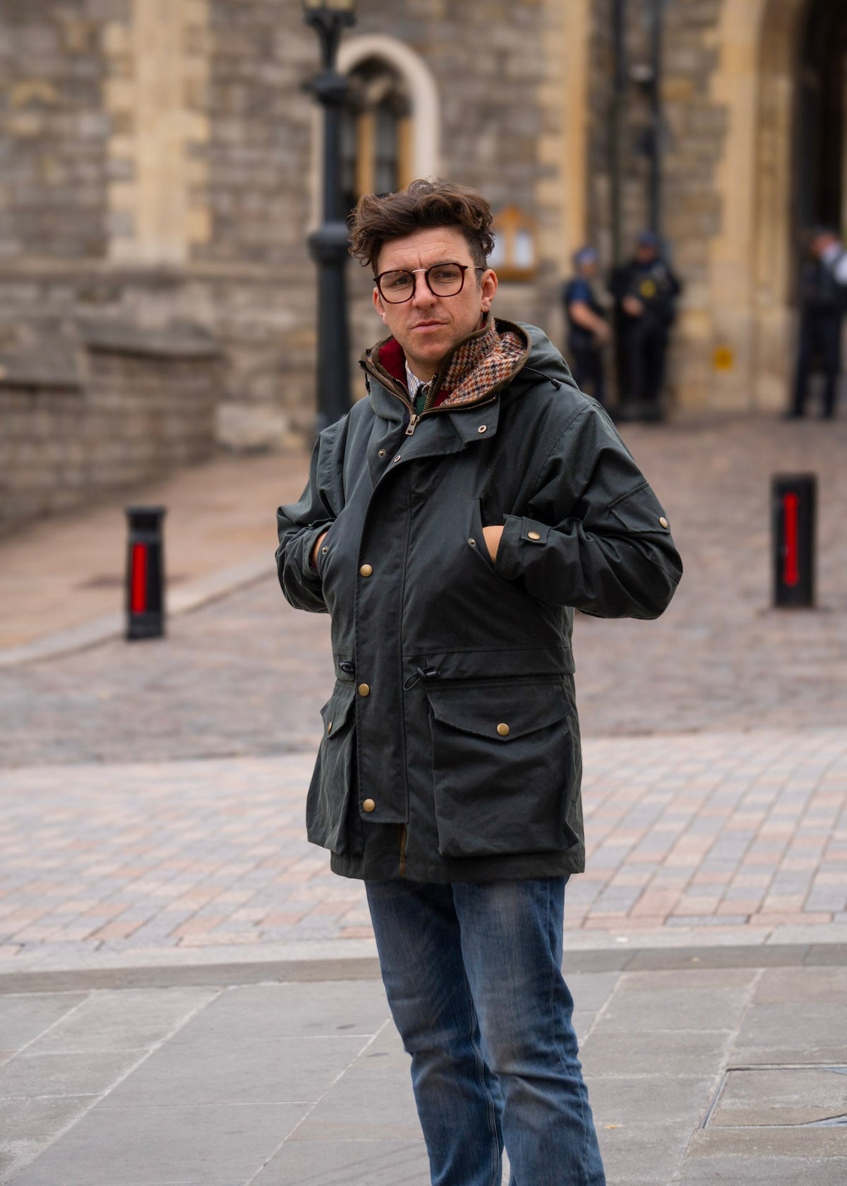 Man wearing a wax jacket and glasses standing in front of a stone building.