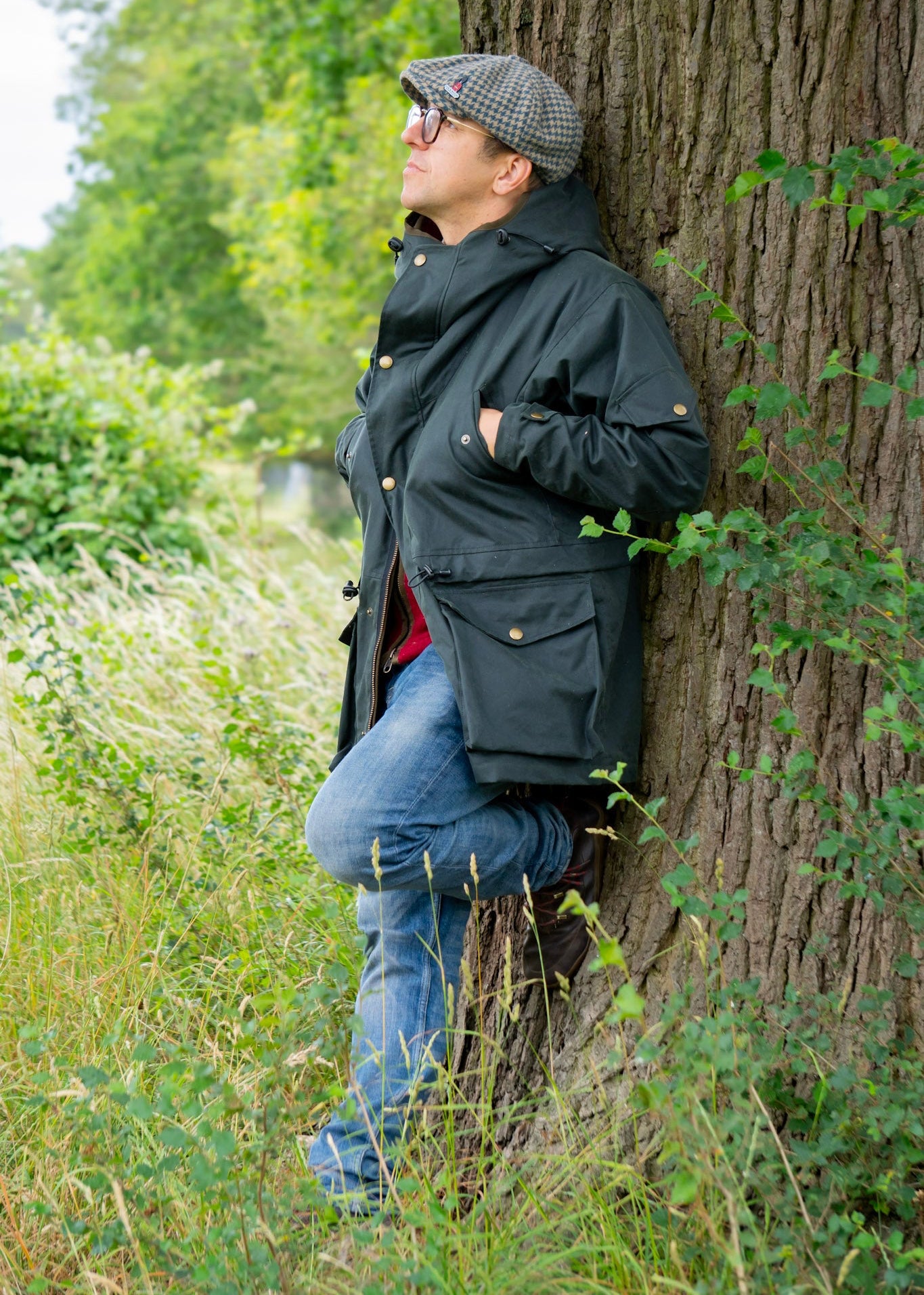 Person leaning against a tree in a natural setting wearing a wax jacket and flat cap