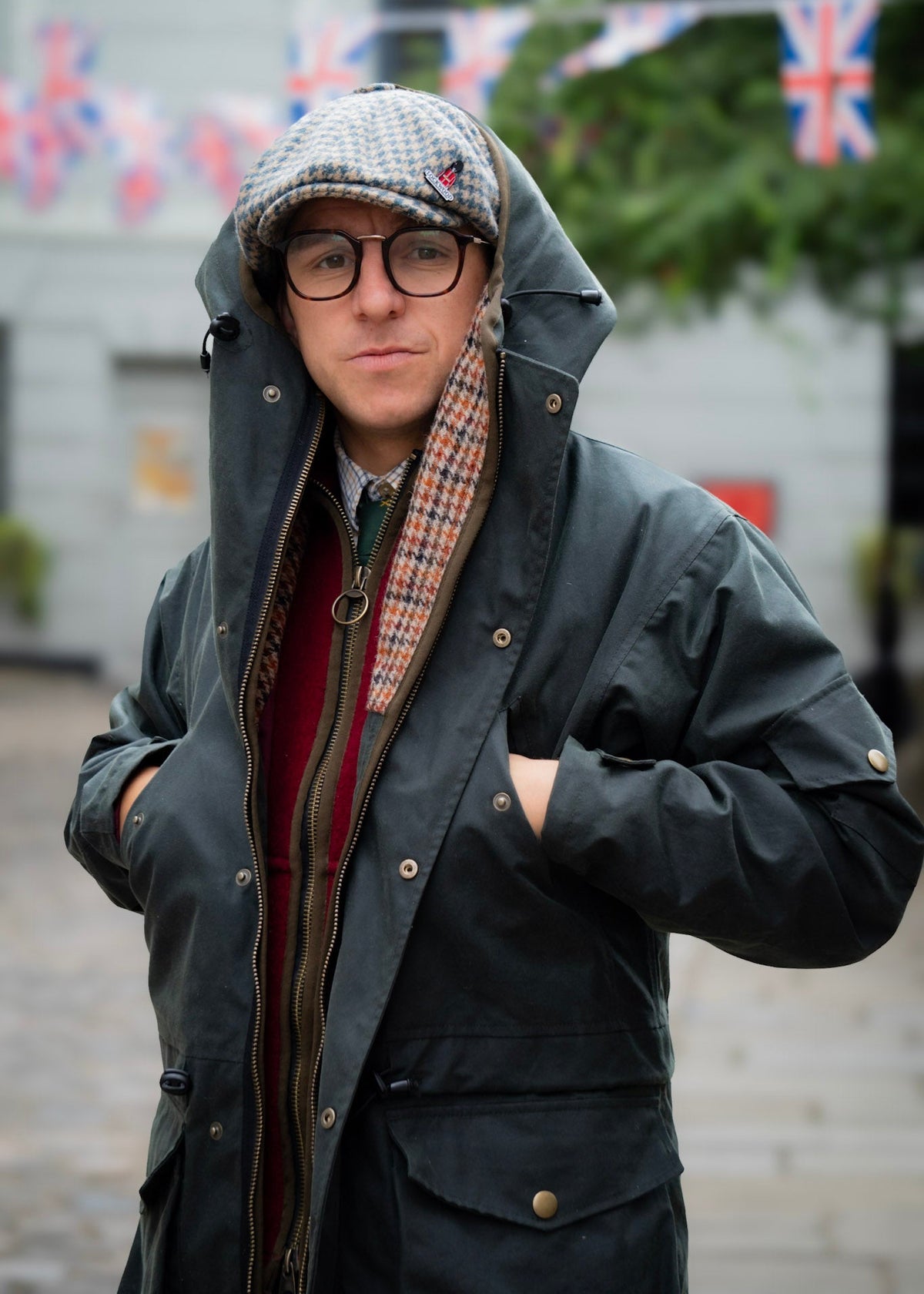 Person wearing a dark wax jacket, red gilet, and glasses with British flags in the background