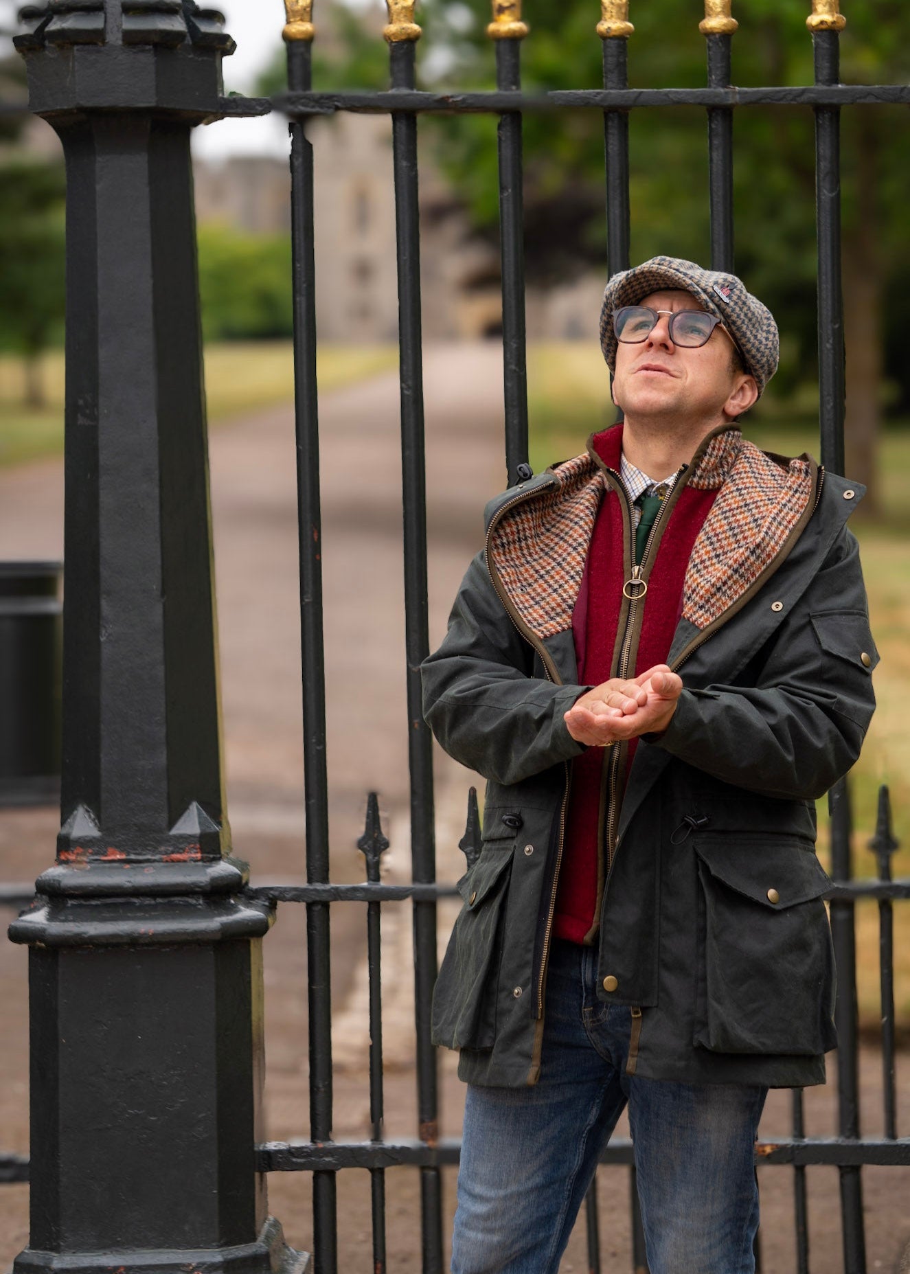 Man standing in front of a black metal gate wearing glasses and a wax jacket with a plaid pattern.