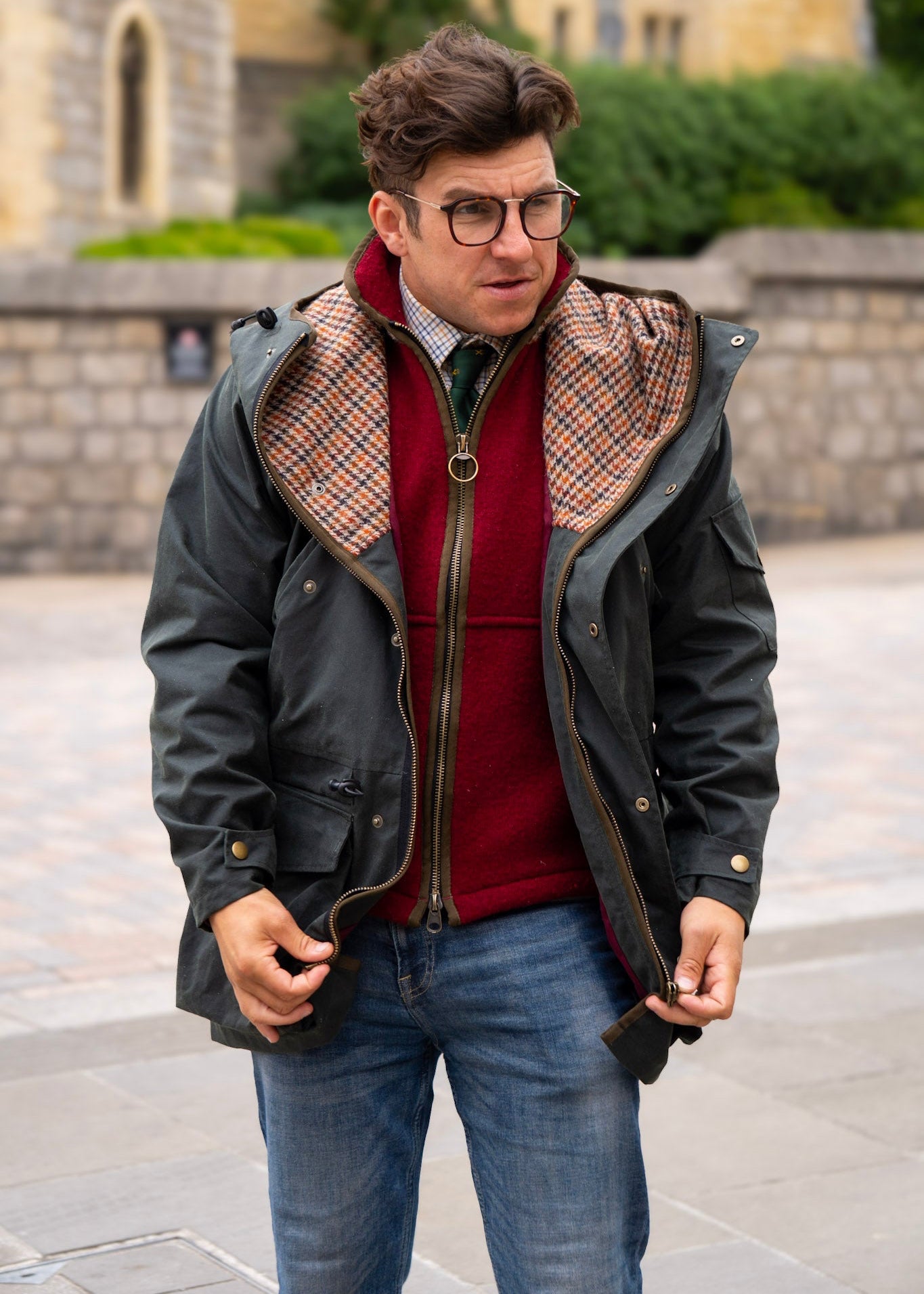Man wearing a dark wax jacket with plaid lining, red gilet, and blue jeans outdoors.