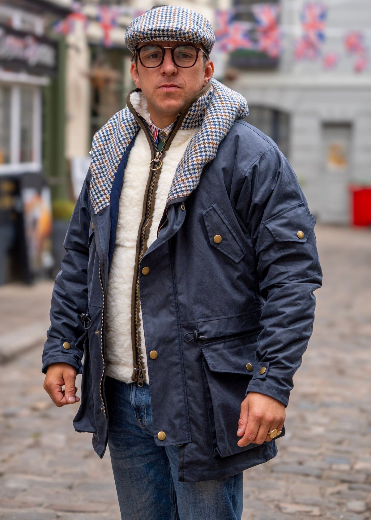Man wearing a navy blue jacket with a patterned interior, glasses, and a cap in an urban setting.
