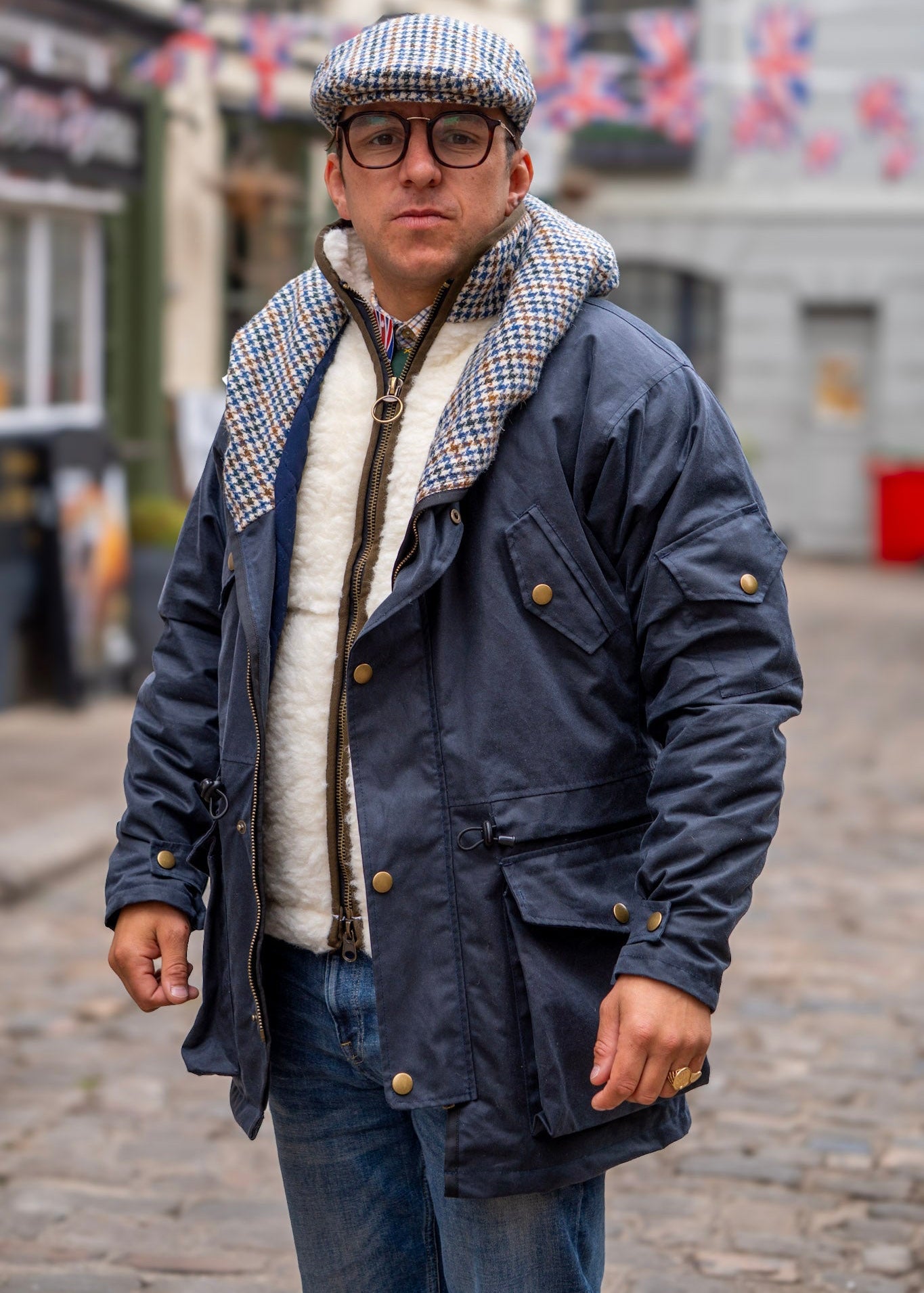 Man wearing a navy blue jacket with a patterned interior, glasses, and a cap in an urban setting.