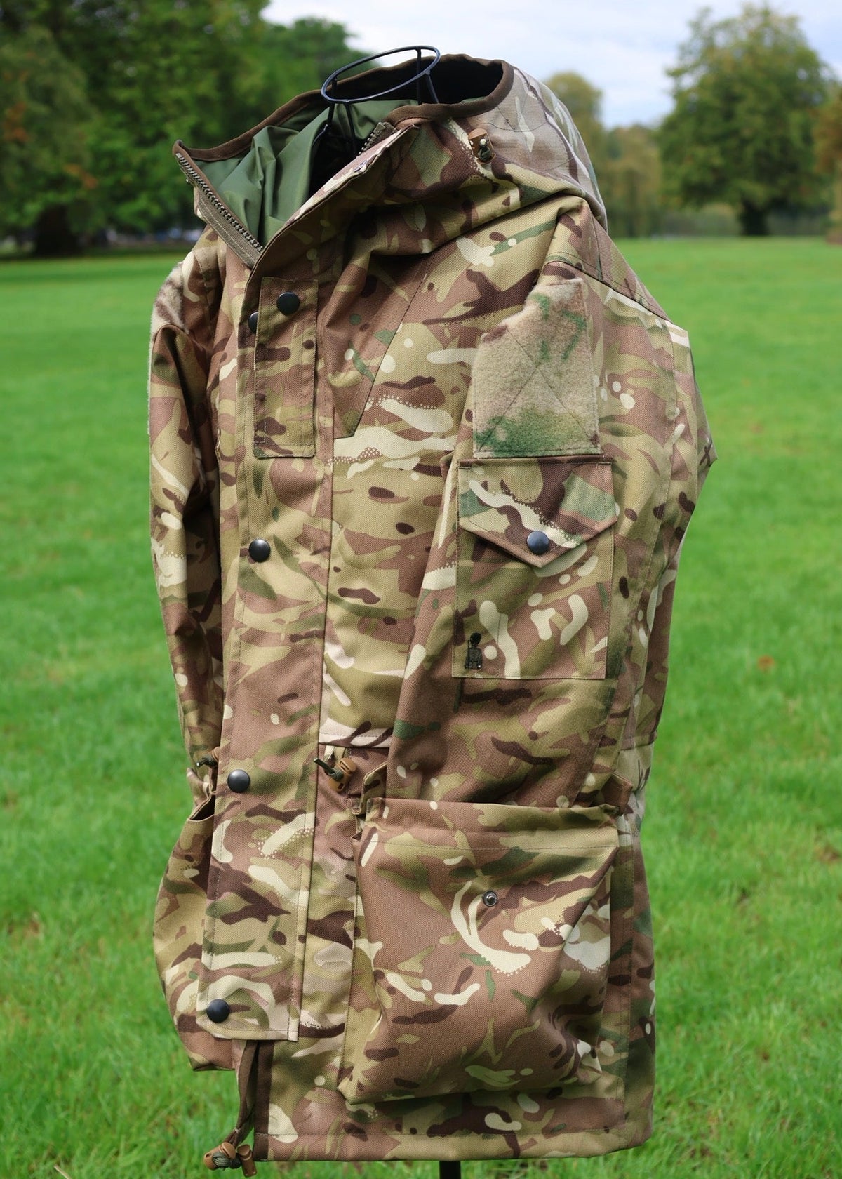 A military-style camouflage jacket displayed on a mannequin outdoors in a grassy park setting.