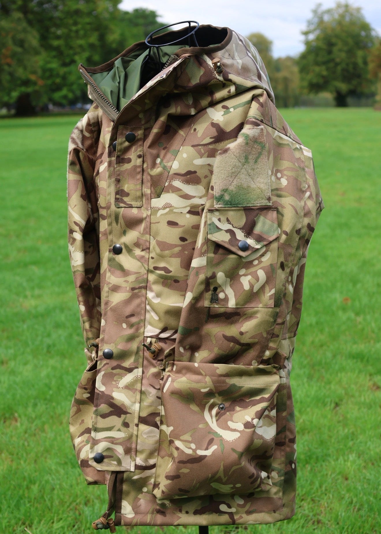 A military-style camouflage jacket displayed on a mannequin outdoors in a grassy park setting.