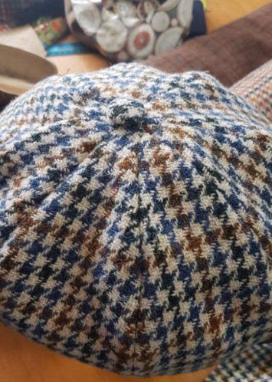 A checkered 8-panel flat cap made of Harris Tweed, displayed on a table with other items.