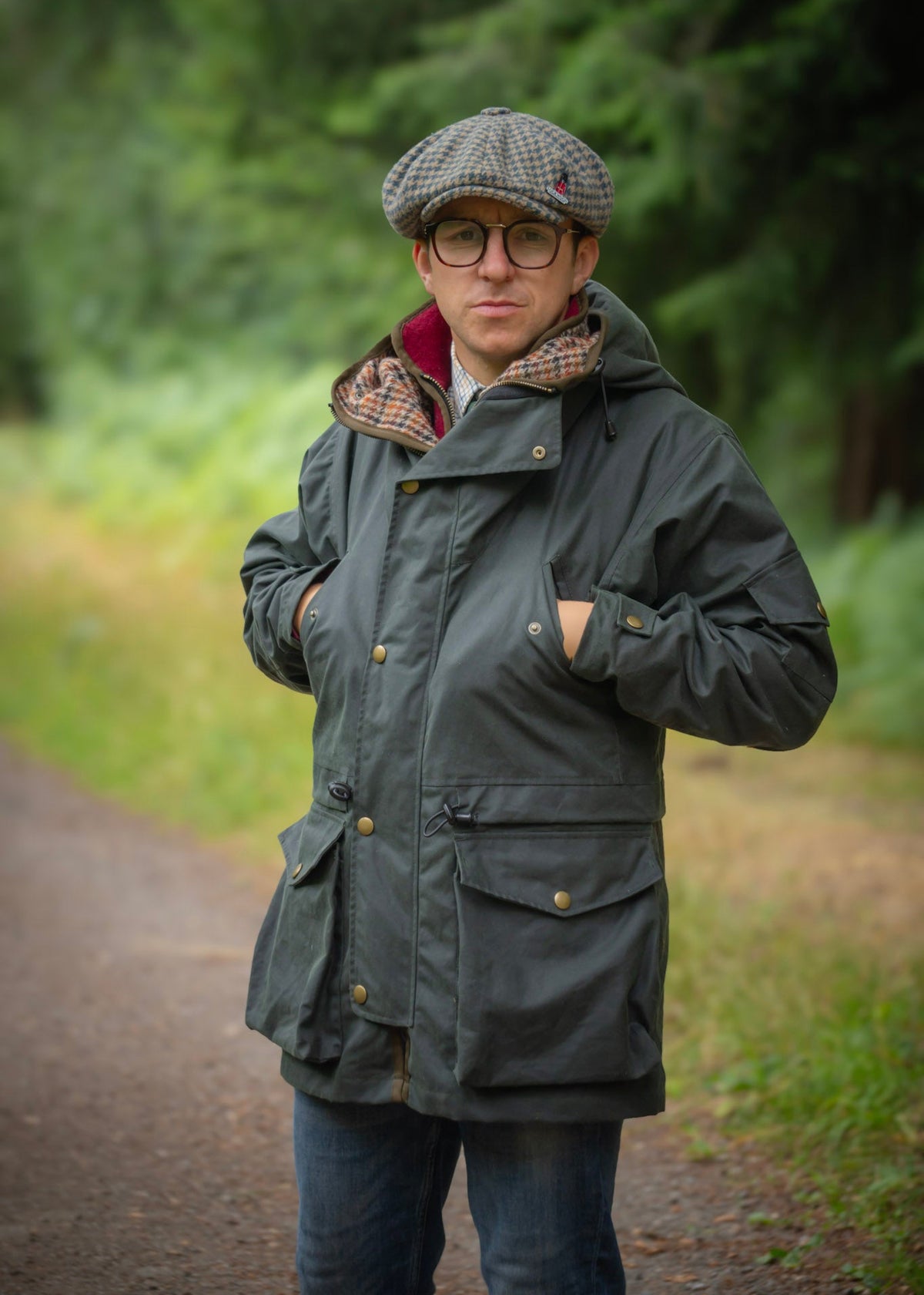 Man wearing a green wax jacket  and flat cap standing on a path with trees in the background