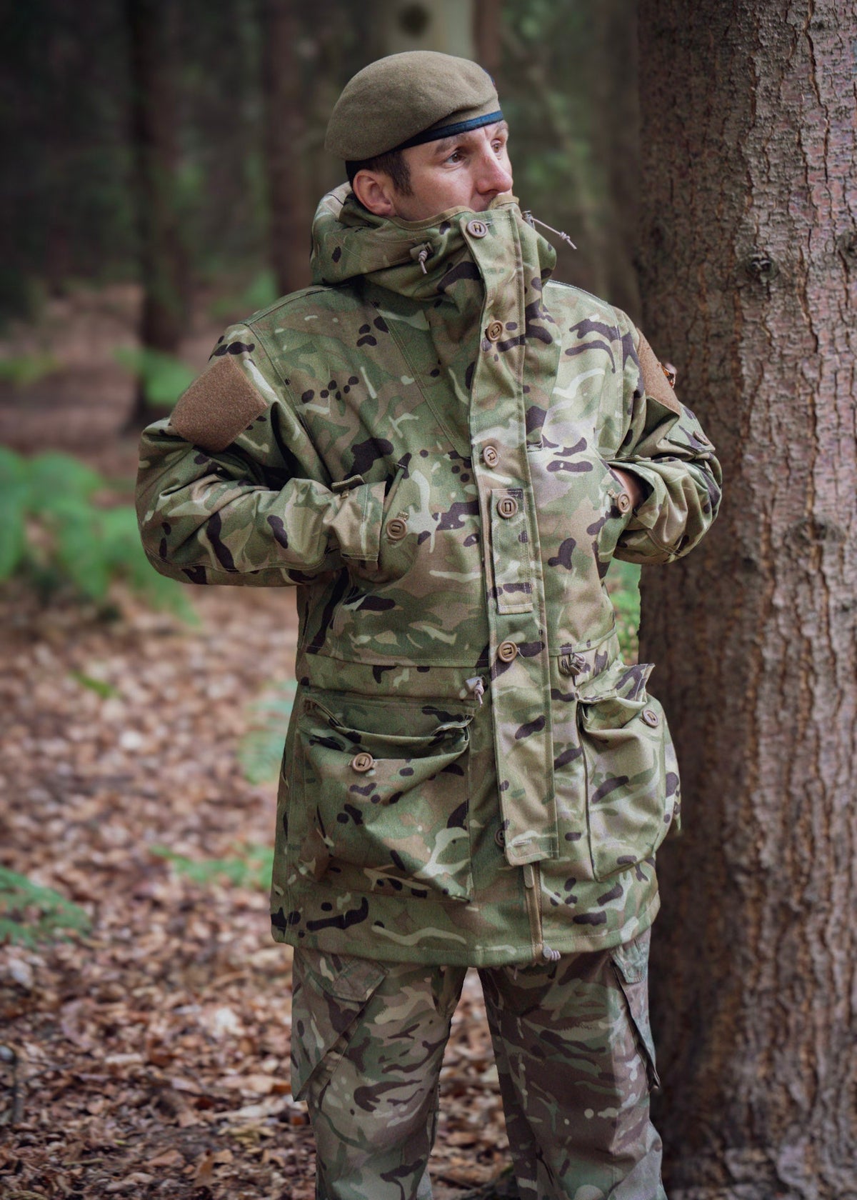 Person in camouflage clothing standing next to a tree in a forest setting