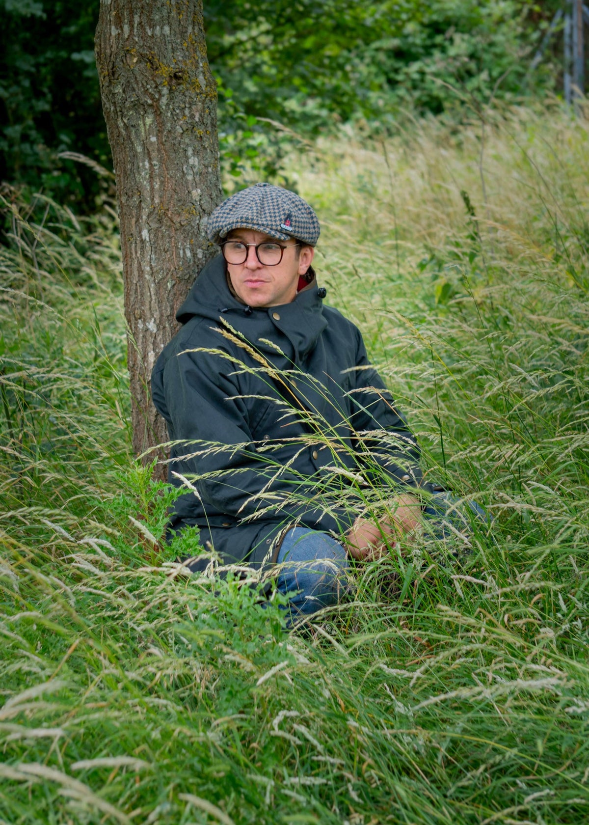 Person sitting in tall grass wearing a wax jacket, cap and glasses, with trees in the background