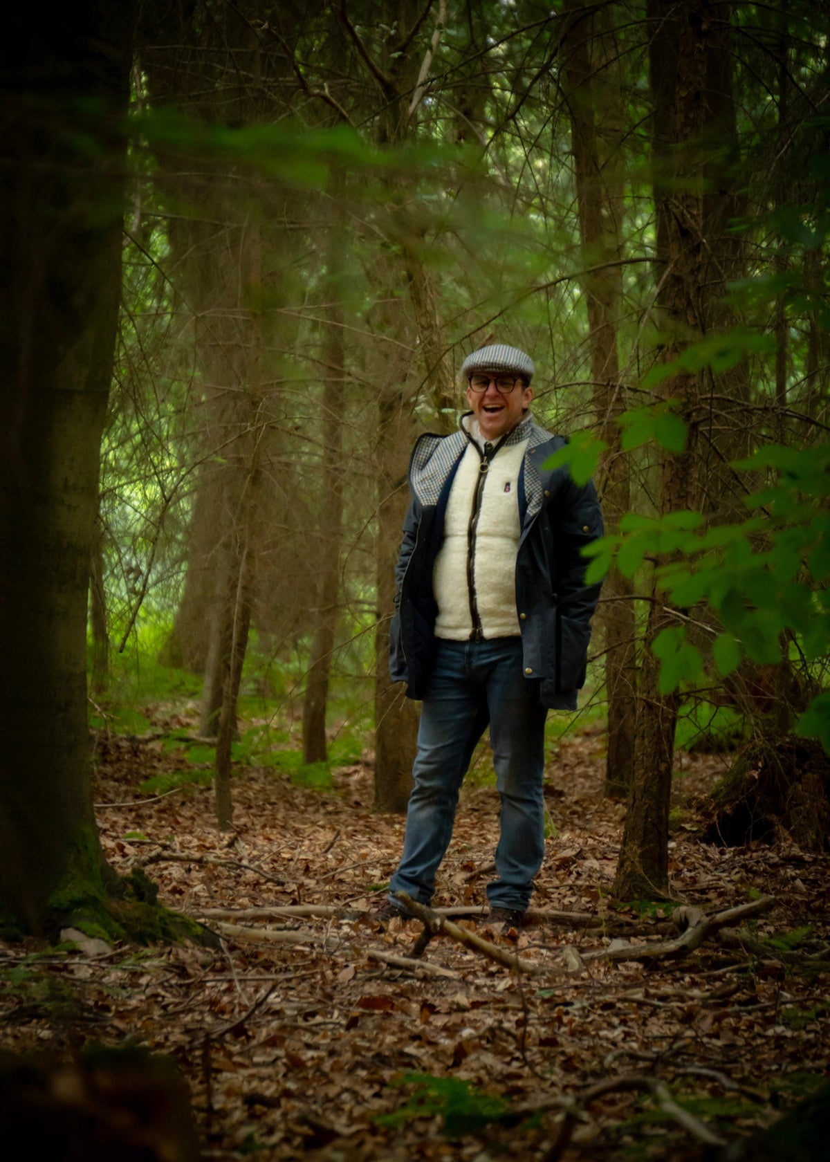 Man standing in a forest with trees and foliage around wearing wax jacket and wool gilet