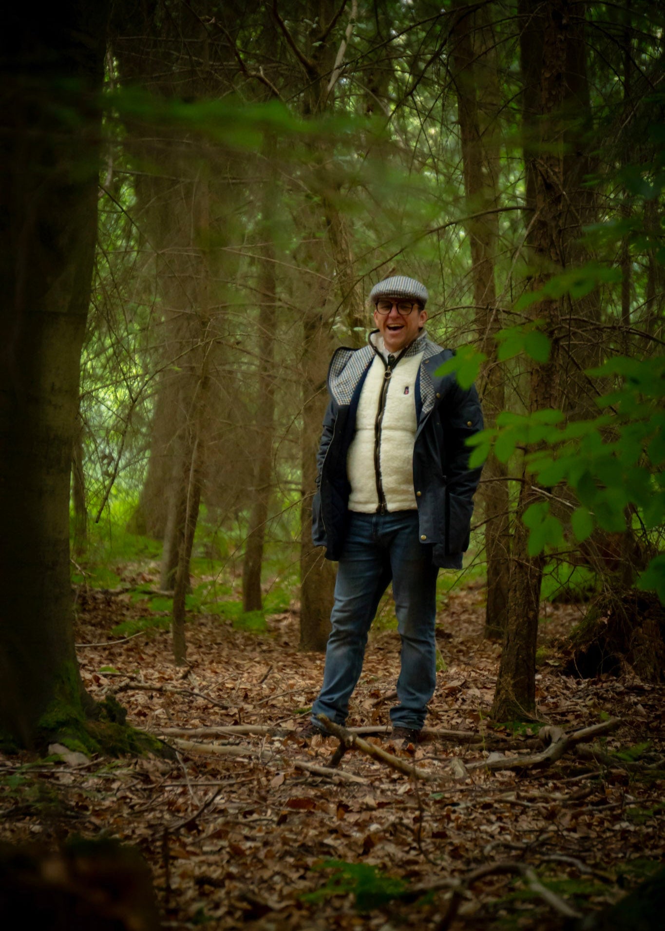 Man standing in a forest with trees and foliage around wearing wax jacket and wool gilet