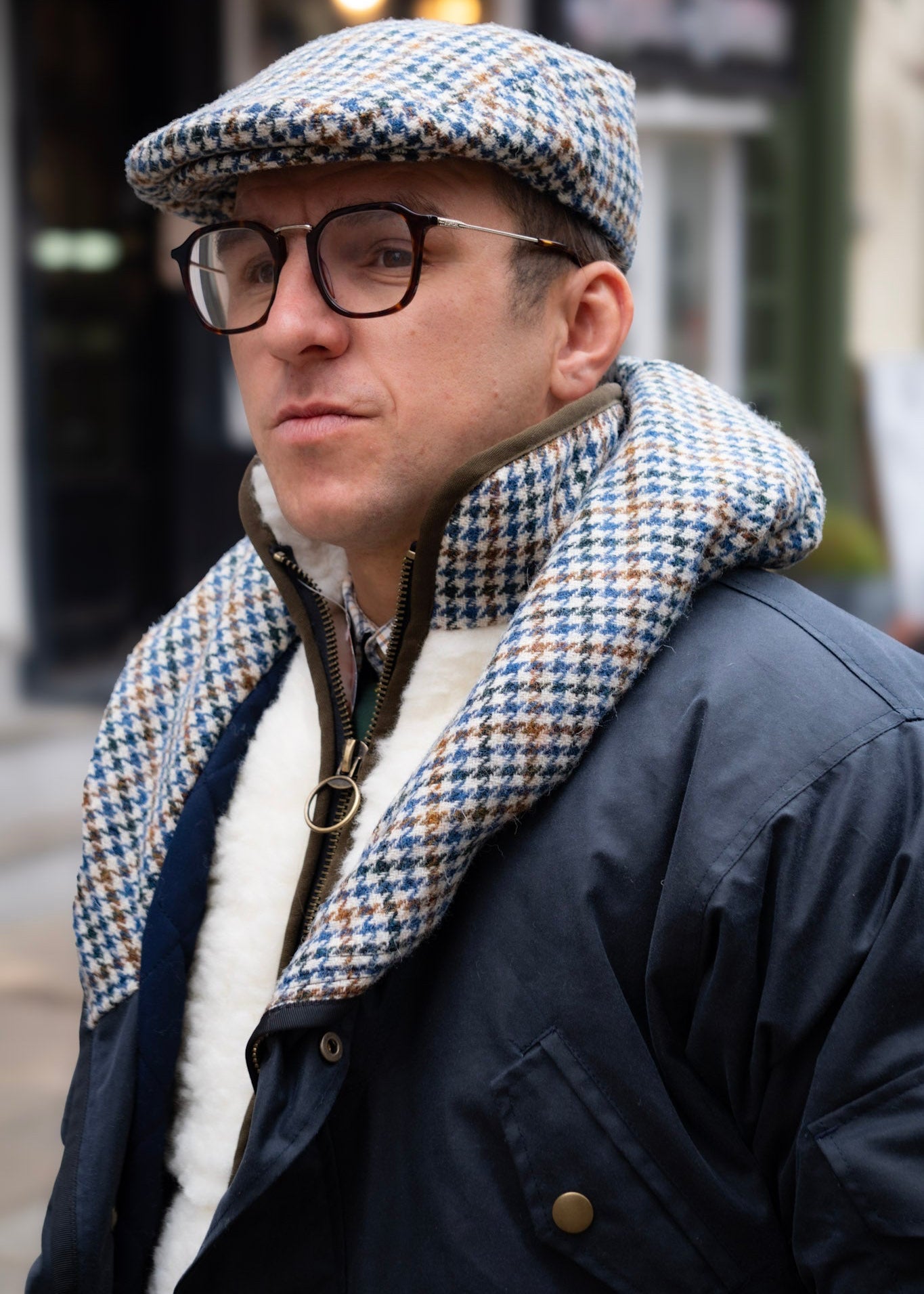 Man wearing a patterned cap, glasses, and a wax jacket in an urban setting