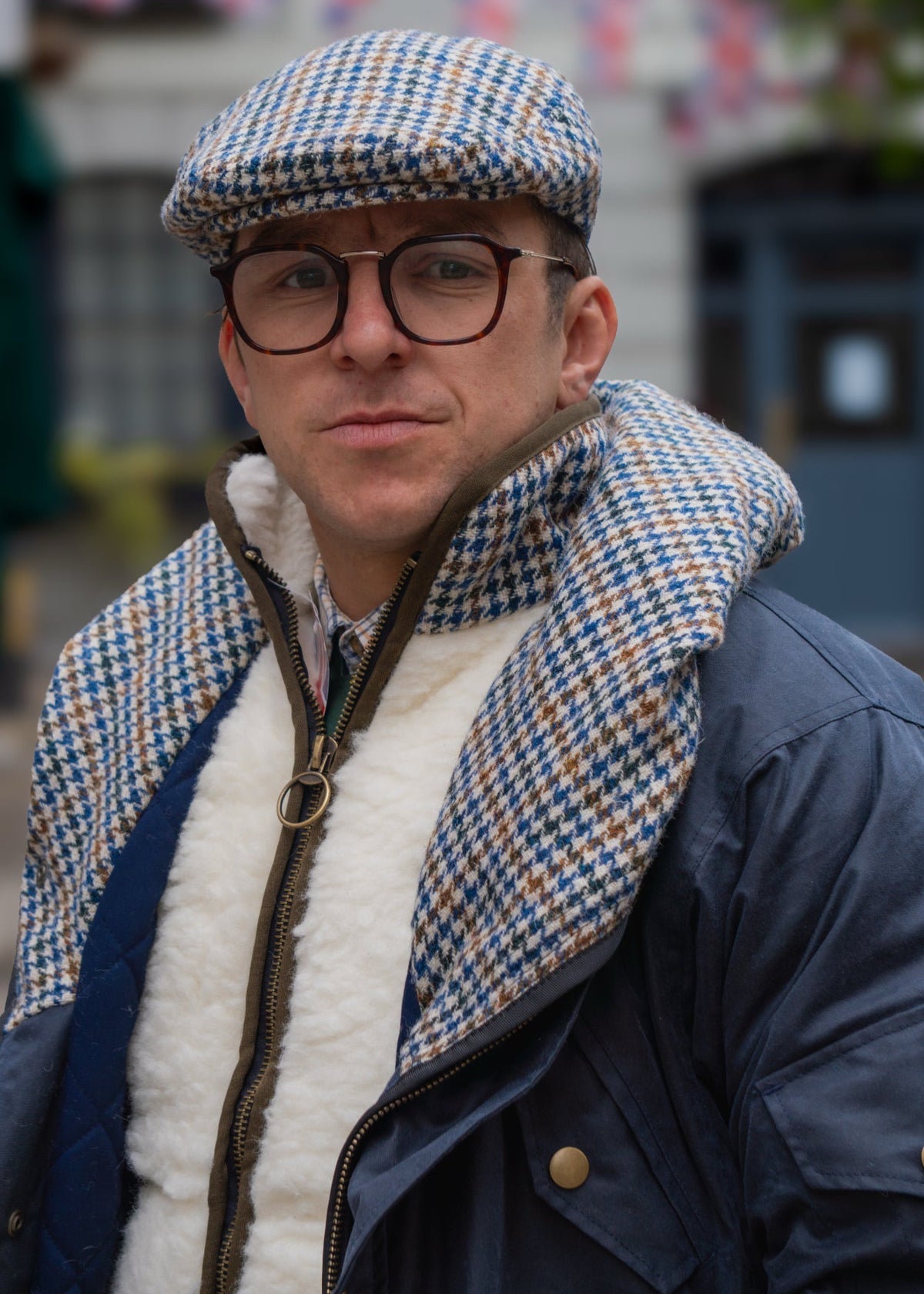 Man wearing a checkered flat cap, glasses, and a dark coat with a white fur gilet