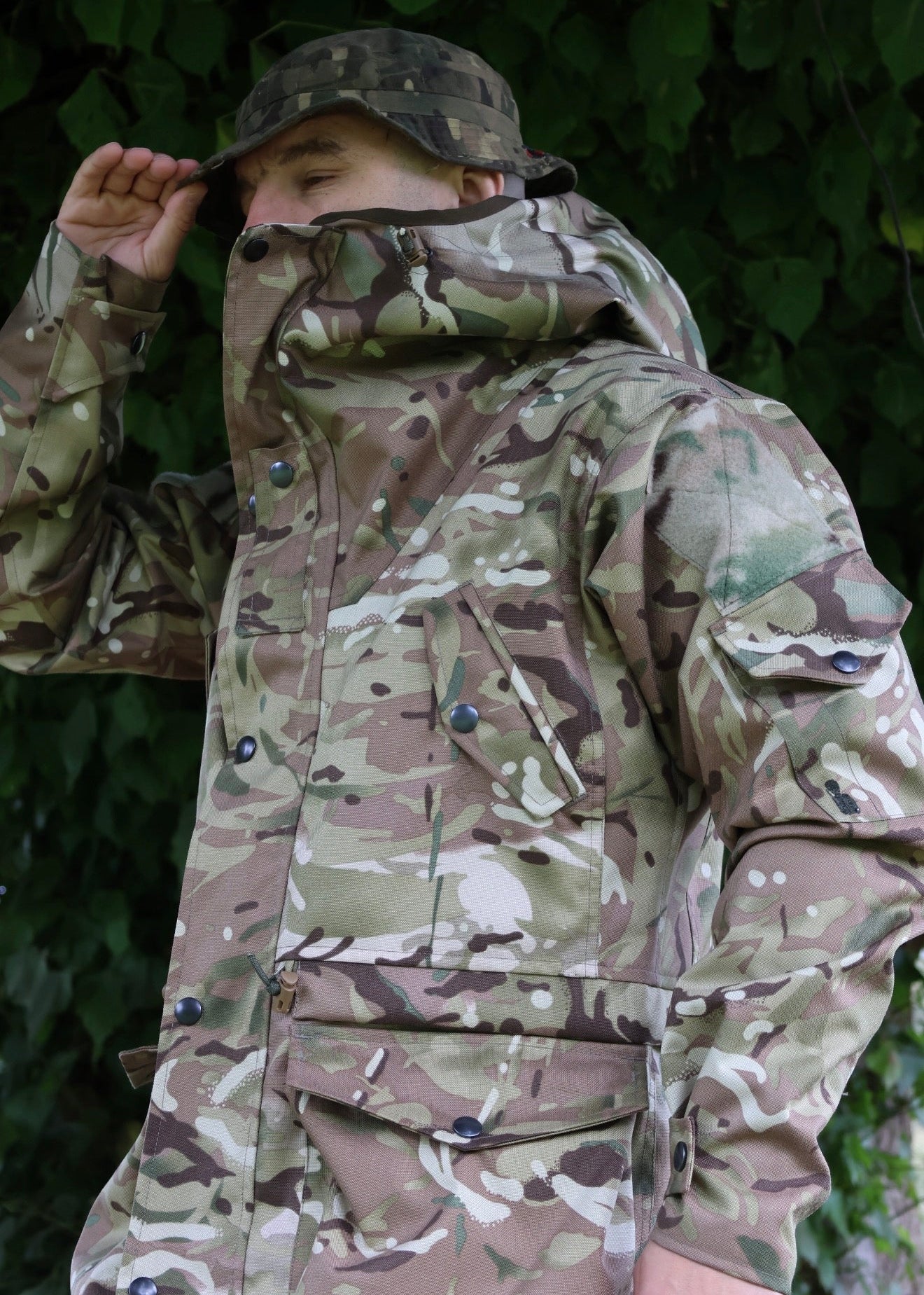 A man wearing a military-style camouflage jacket and matching camouflage cap, standing outdoors in front of a leafy green background.
