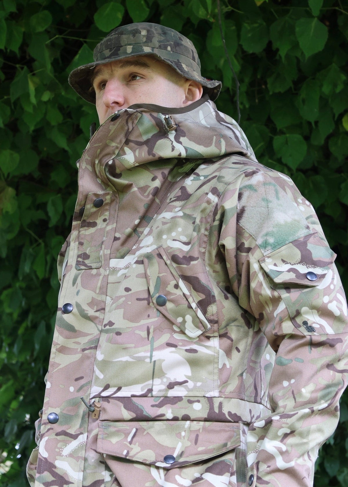 A man wearing a camouflage hooded jacket and matching camouflage bucket hat, standing outdoors in front of a background of green leaves.