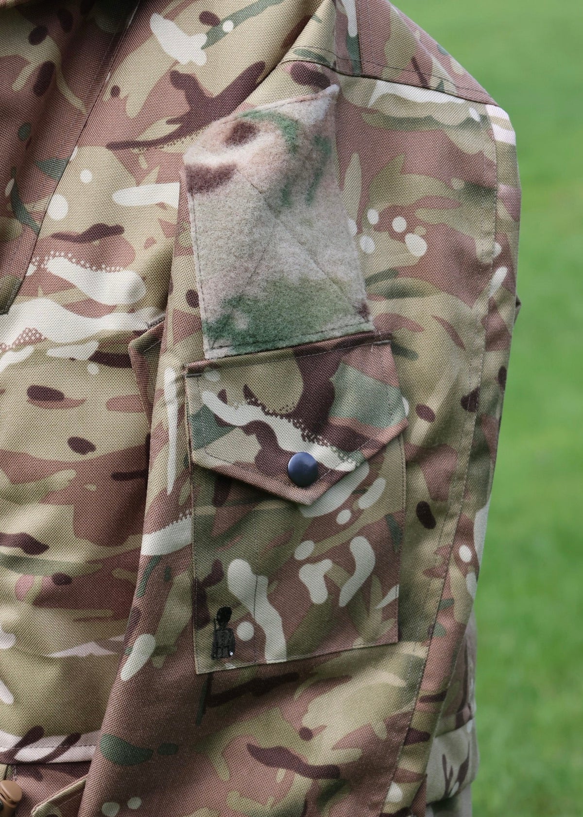 Close-up of a camouflage military-style jacket with a flap pocket and a green camouflage patch on the shoulder, set against a blurred green grass background.