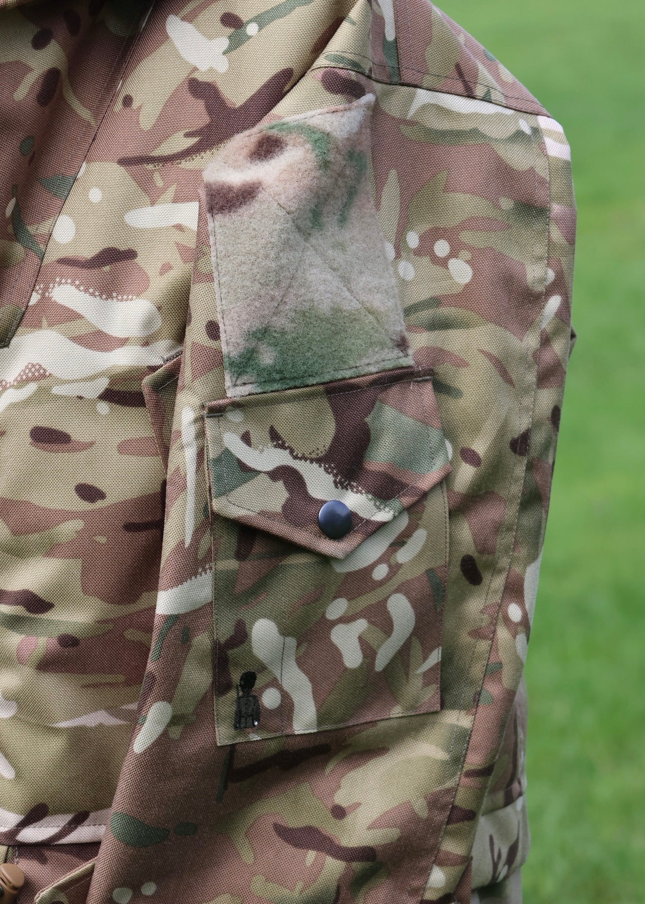 Close-up of a camouflage military-style jacket with a flap pocket and a green camouflage patch on the shoulder, set against a blurred green grass background.
