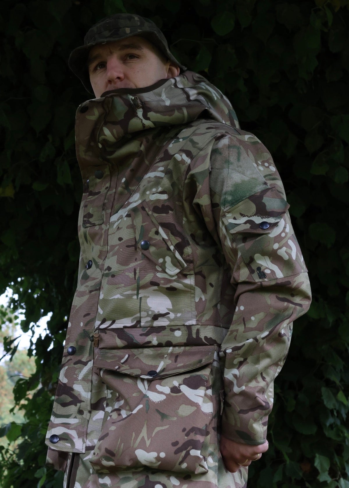 A person wearing a camouflage hooded jacket and matching cap, photographed outdoors against a dark leafy background.