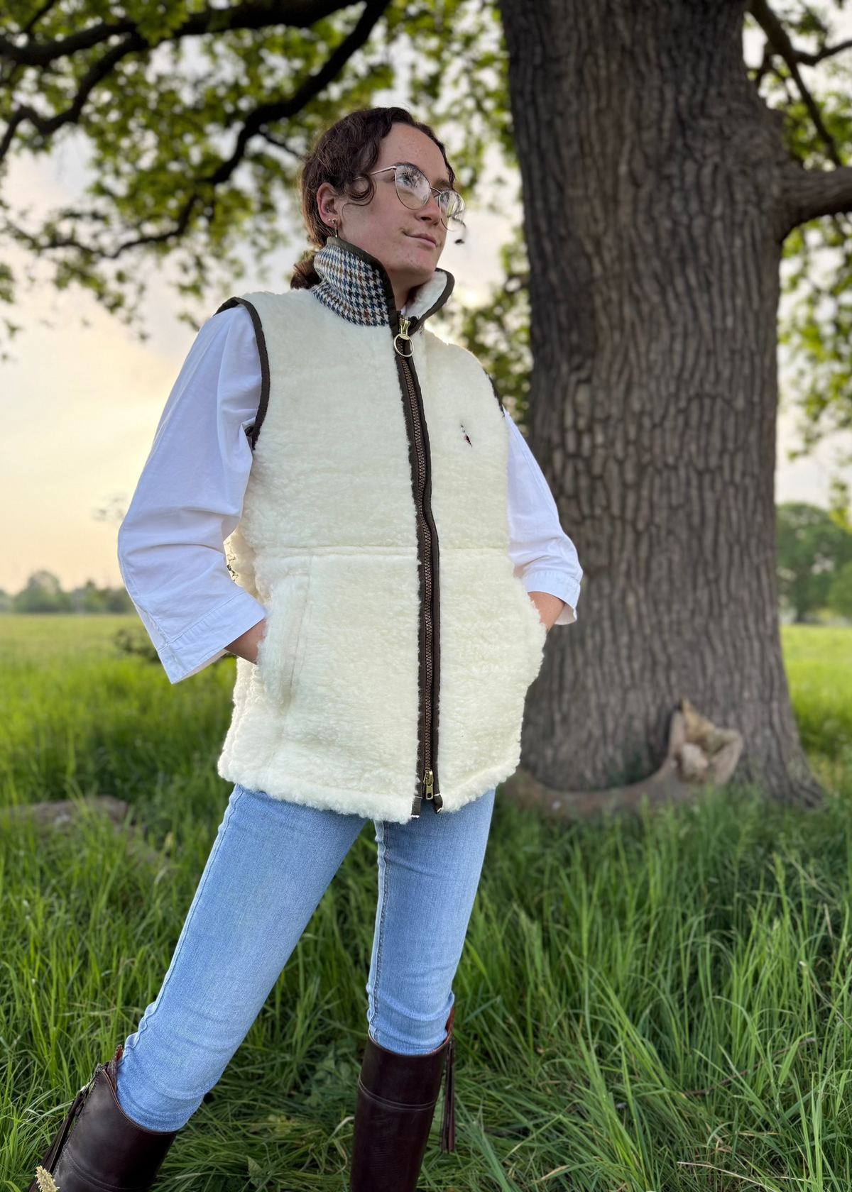 A person standing outdoors wearing a beige-colored lambswool gilet with a Harris Tweed collar, blue jeans, and black boots.