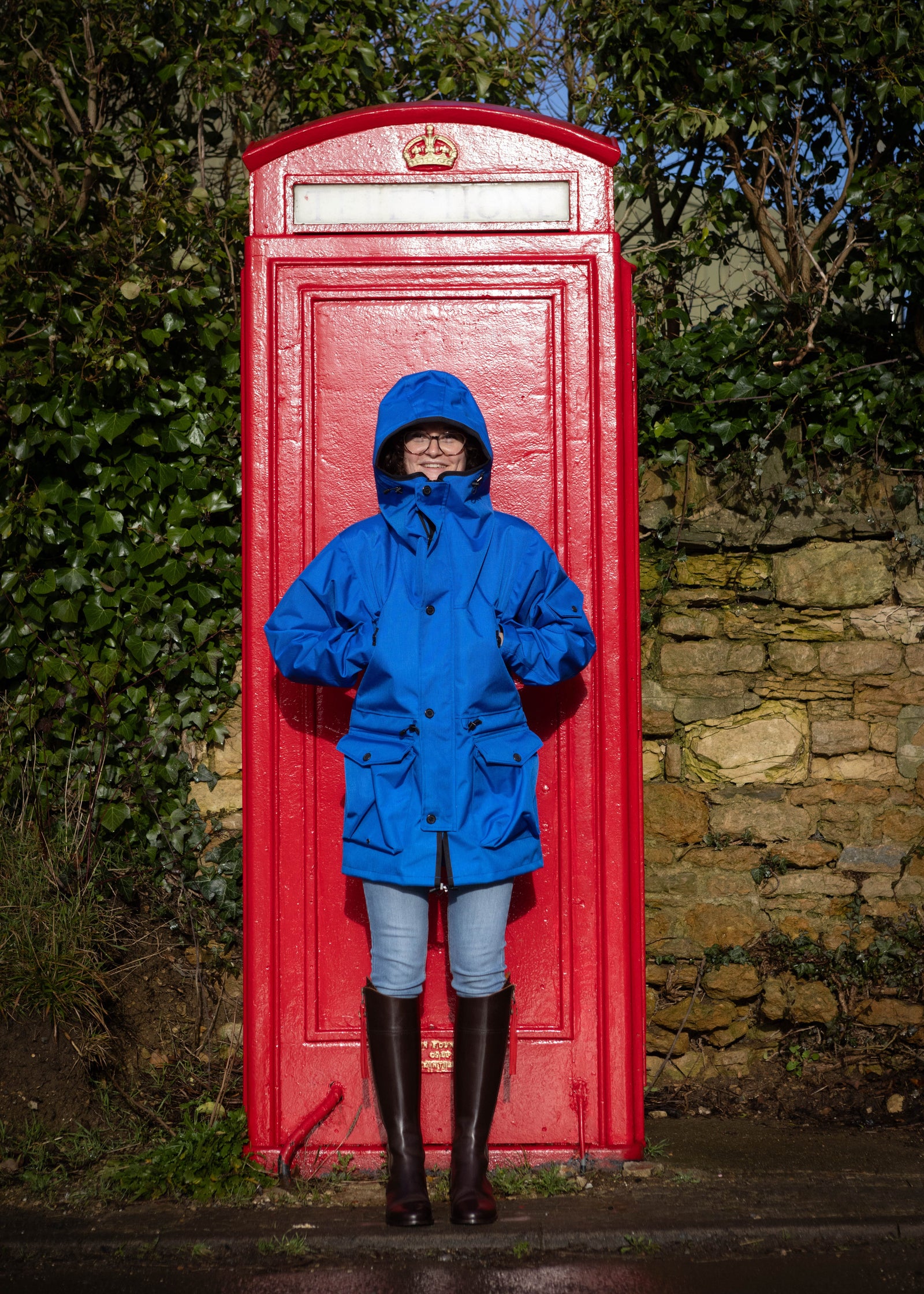 A person wearing a royal blue outdoor smock with an oversized hood standing in front of a red telephone box.