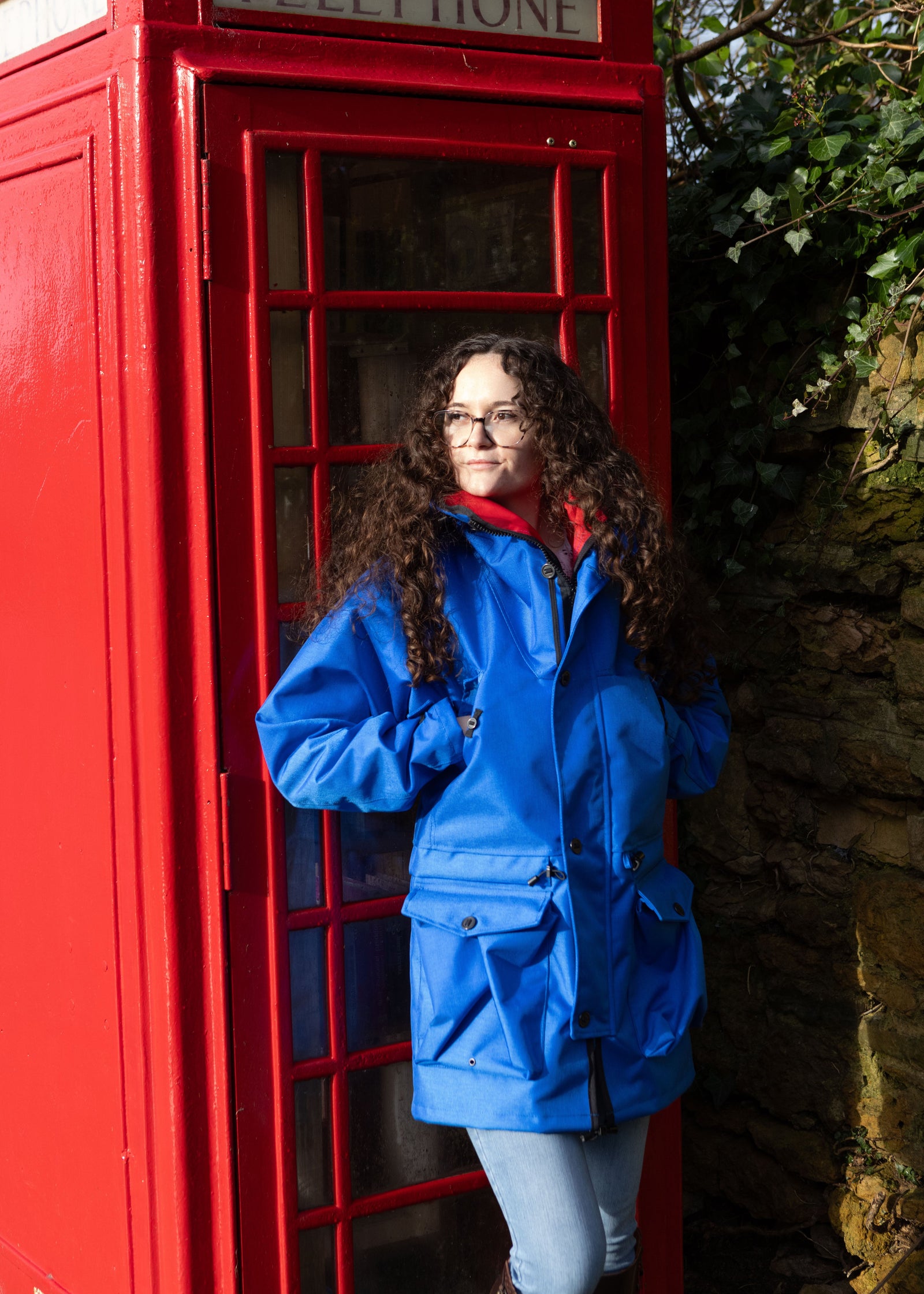 woman wearing a waterproof jacket in royal blue whilst leaning on a British telephone box